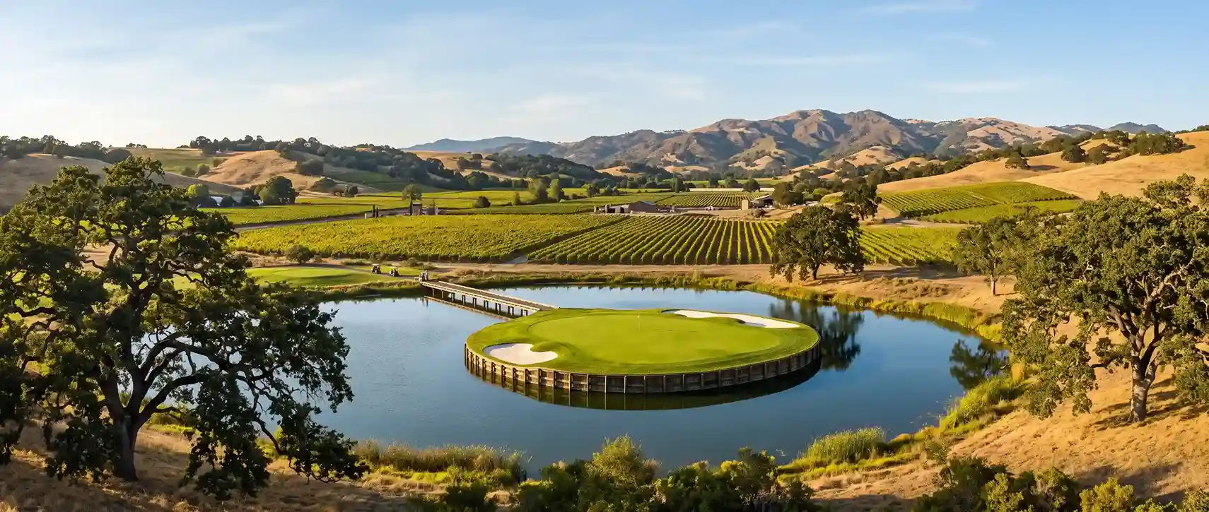 Panoramic view of a circular island golf green surrounded by a lake, footbridge, lush vineyards, and rolling hills.
