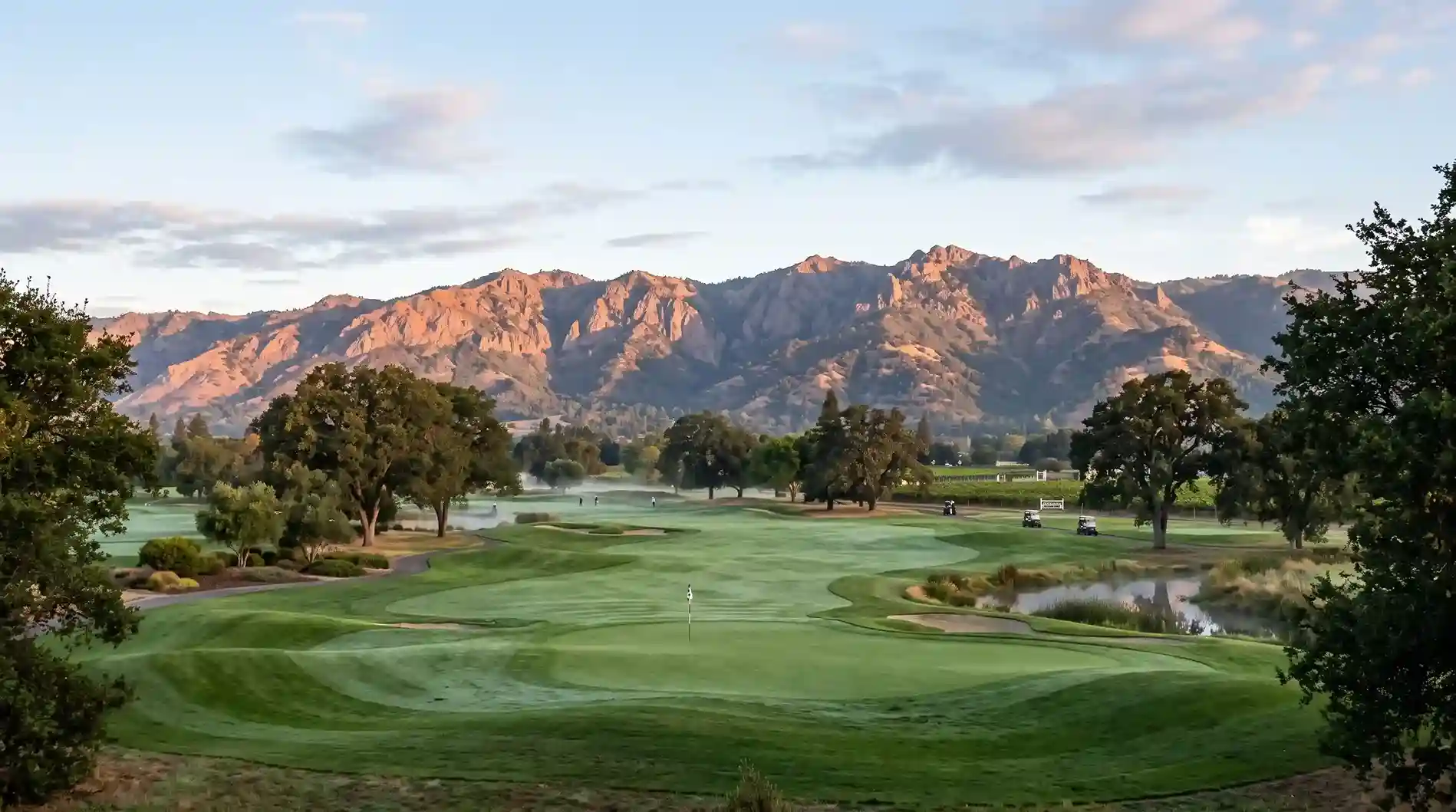 Panoramic view of a lush green golf course fairway and putting green framed by trees and a scenic mountain range glowing in the sunlight.