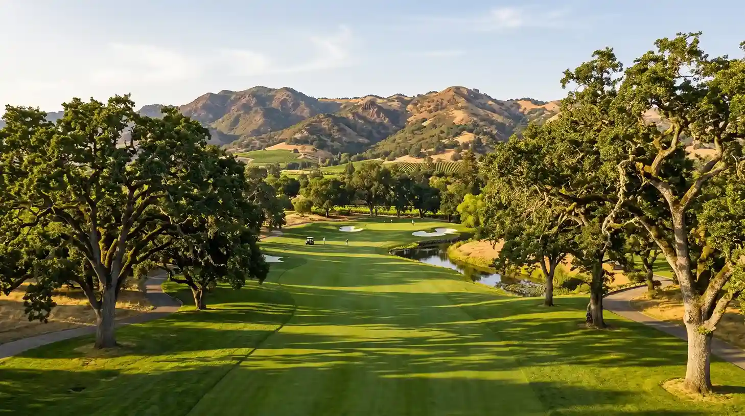 Aerial view of a lush green golf course fairway framed by large oak trees, a water hazard, and sunlit rolling mountains in Napa Valley.